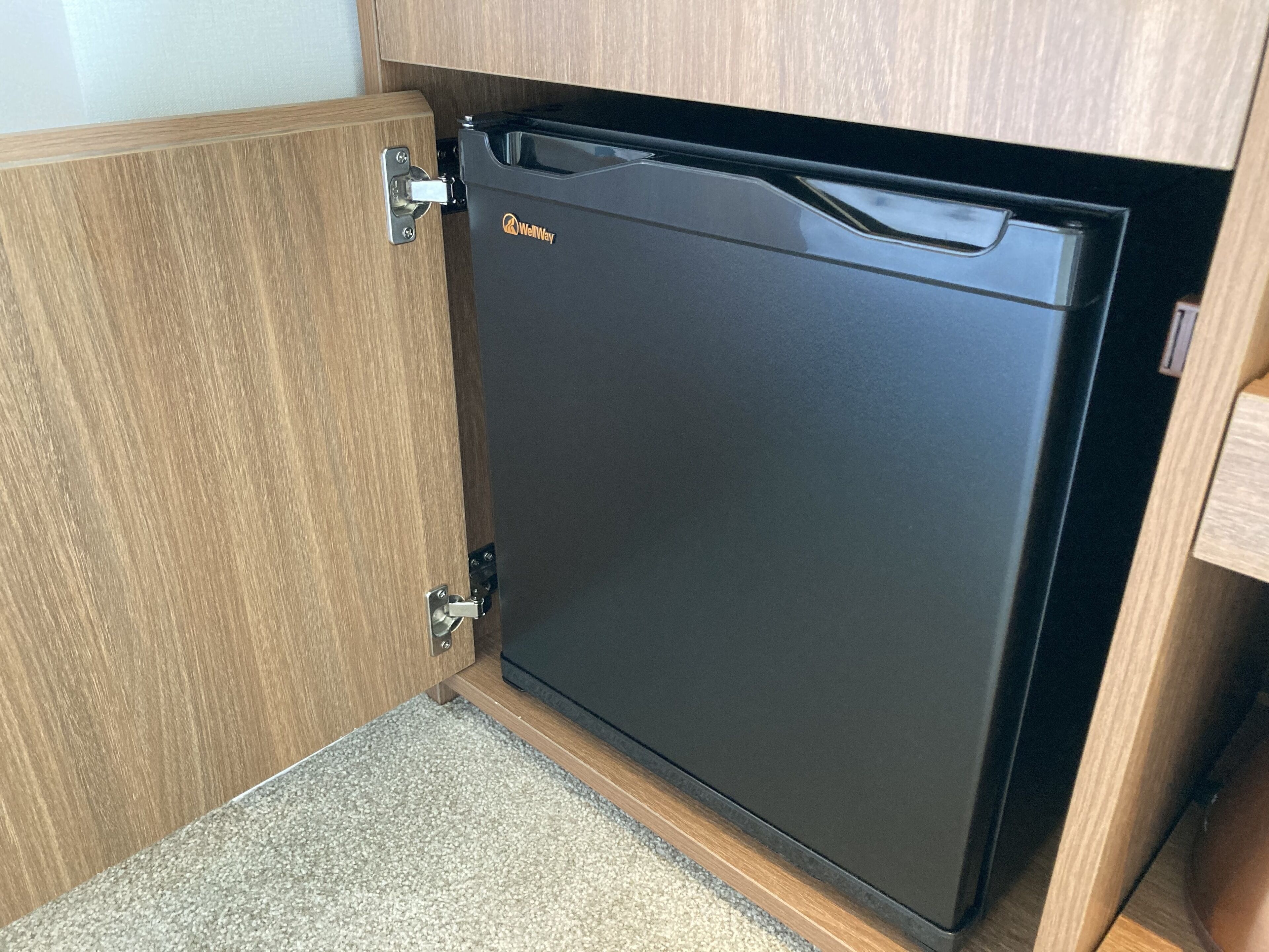 A compact black refrigerator with a WellWay logo, installed under a wooden cabinet in a kitchen setting.