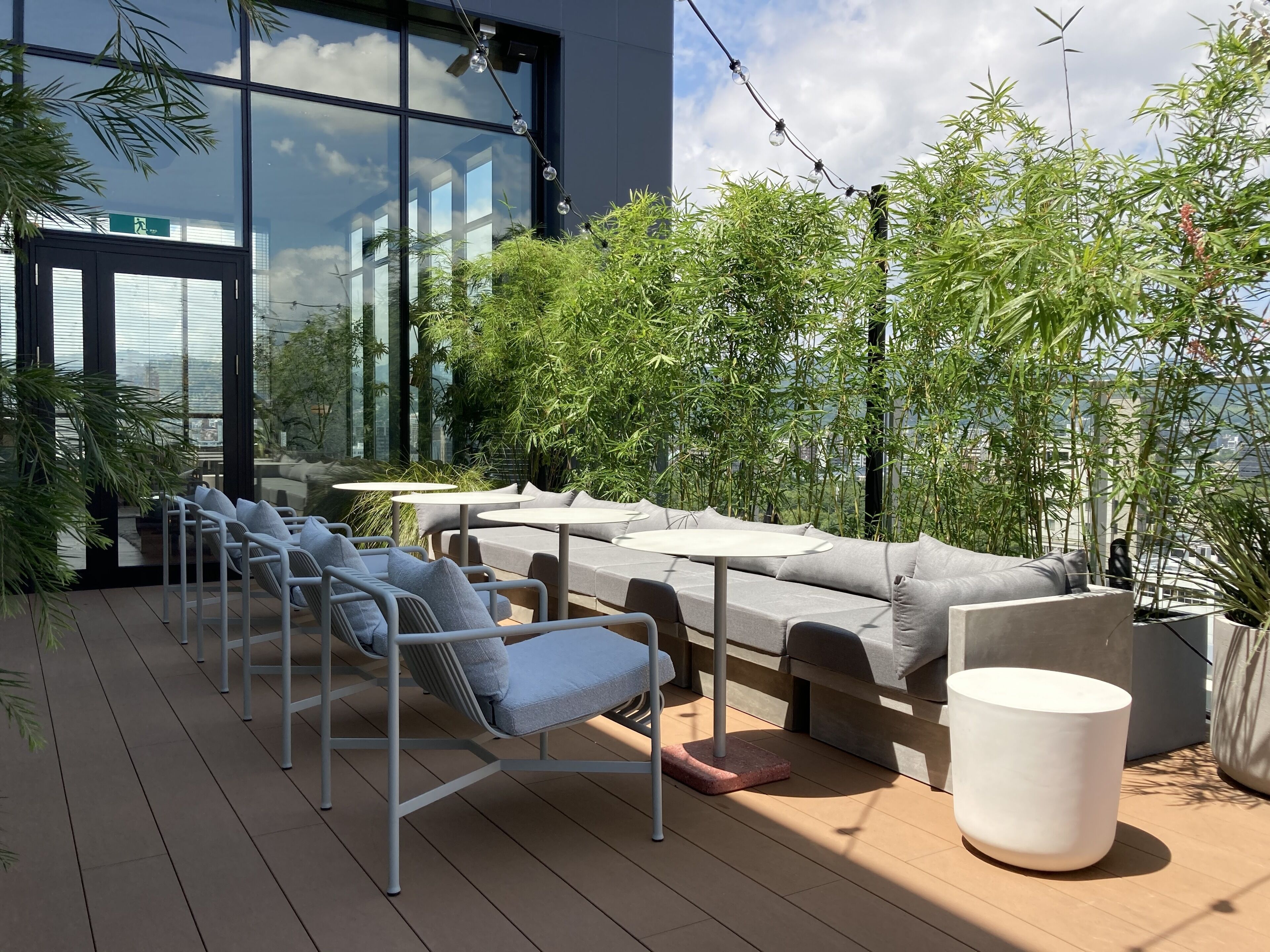 Un área de estar exterior moderna con un sofá y sillones, rodeada de plantas de bambú y vistas a la ciudad.