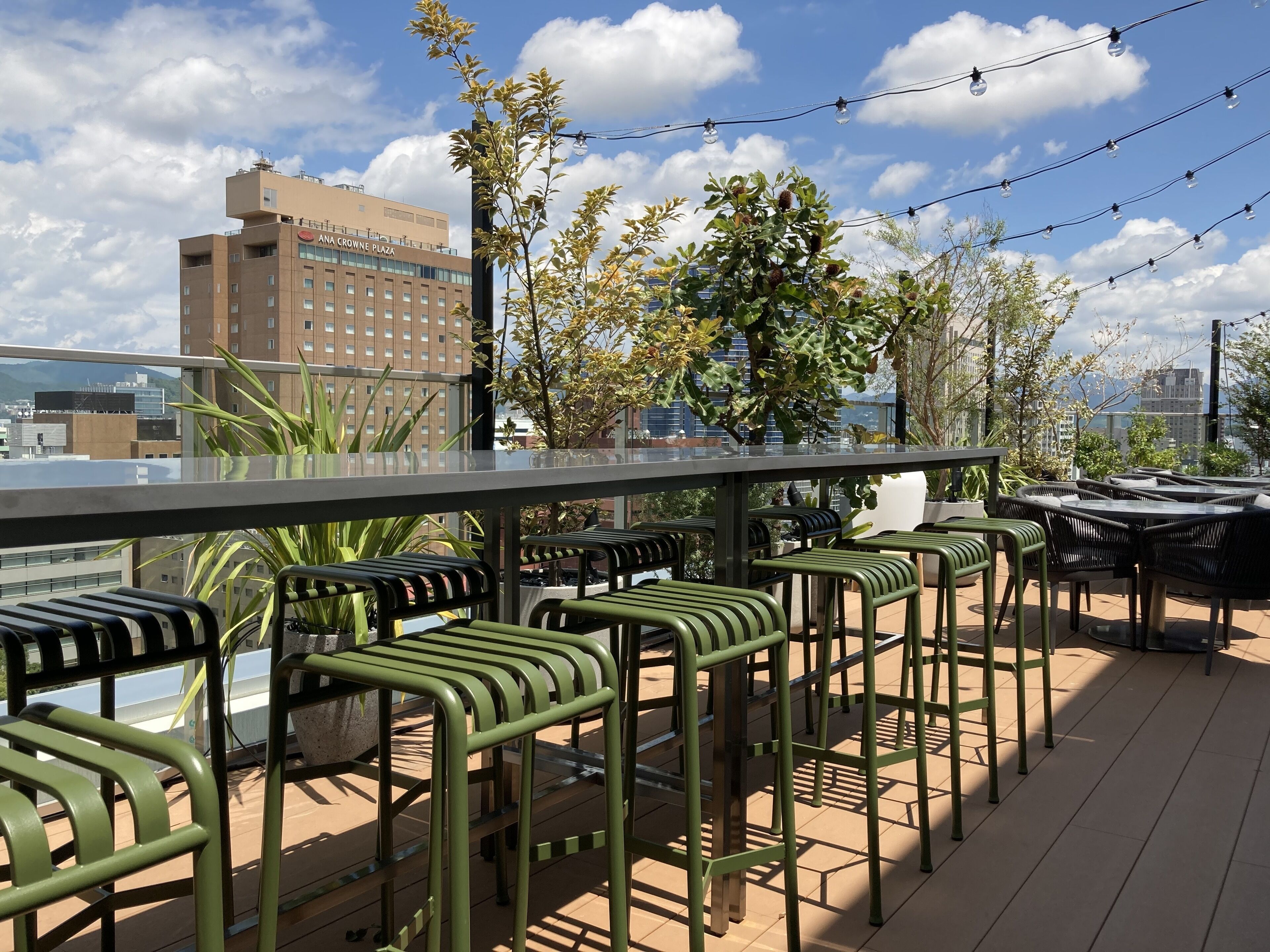 Un bar en la azotea con mesas al aire libre, vistas despejadas del horizonte de la ciudad y un edificio con el rótulo "Crown Plaza" al fondo.