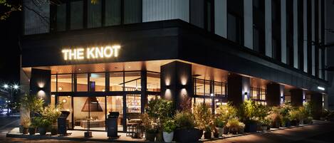Fachada moderna de un hotel con un llamativo letrero iluminado que dice "THE KNOT".
