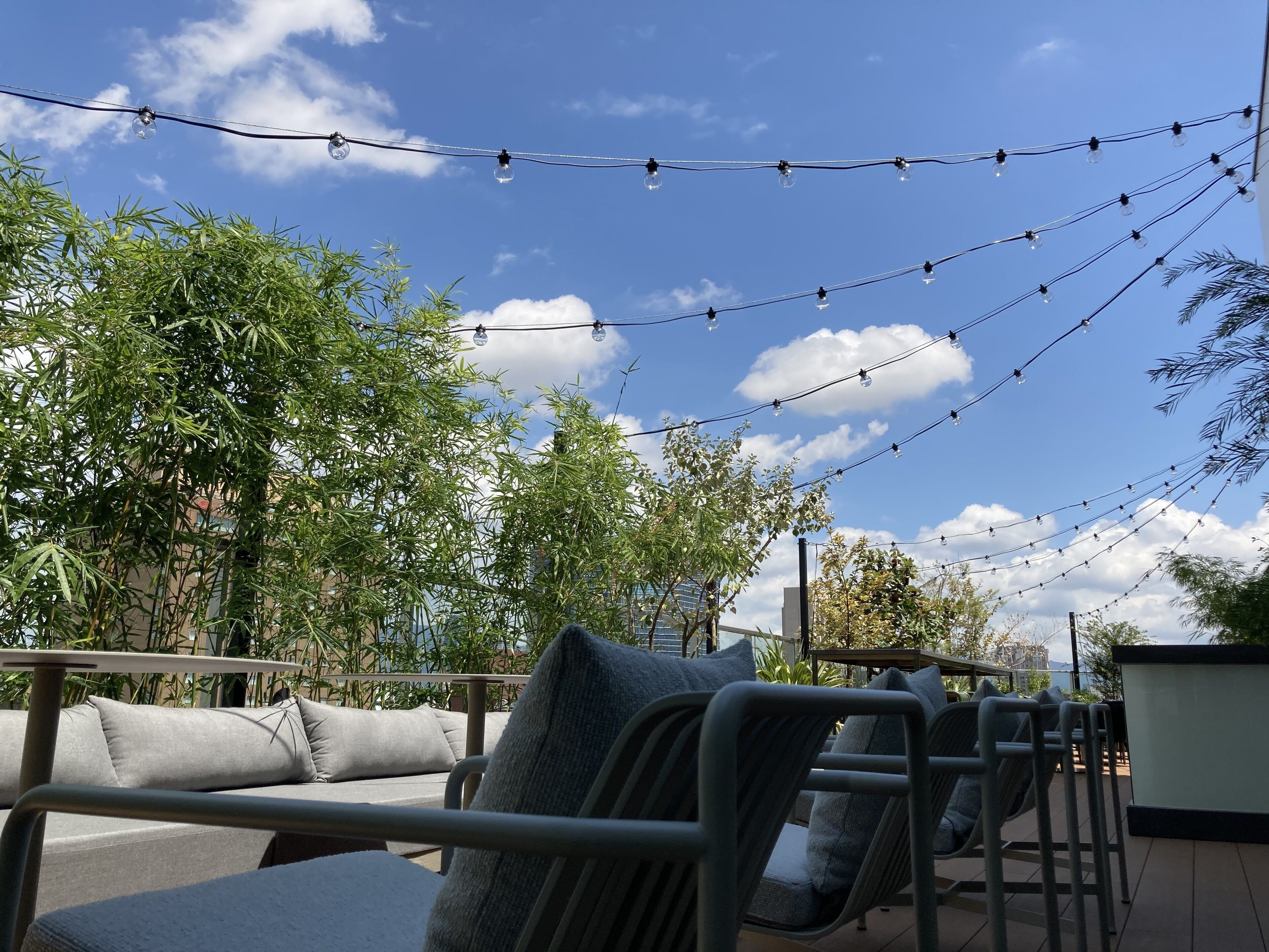Terrasse mit Metallstühlen und Kissen, Lichterketten und Bambuspflanzen unter blauem Himmel.