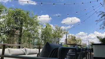 Outdoor seating with metal chairs and cushions, string lights, and bamboo trees under a clear sky.