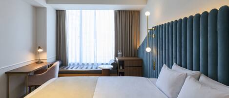 A modern hotel room with a large bed, a desk, a chair, a small table, and a window with curtains.