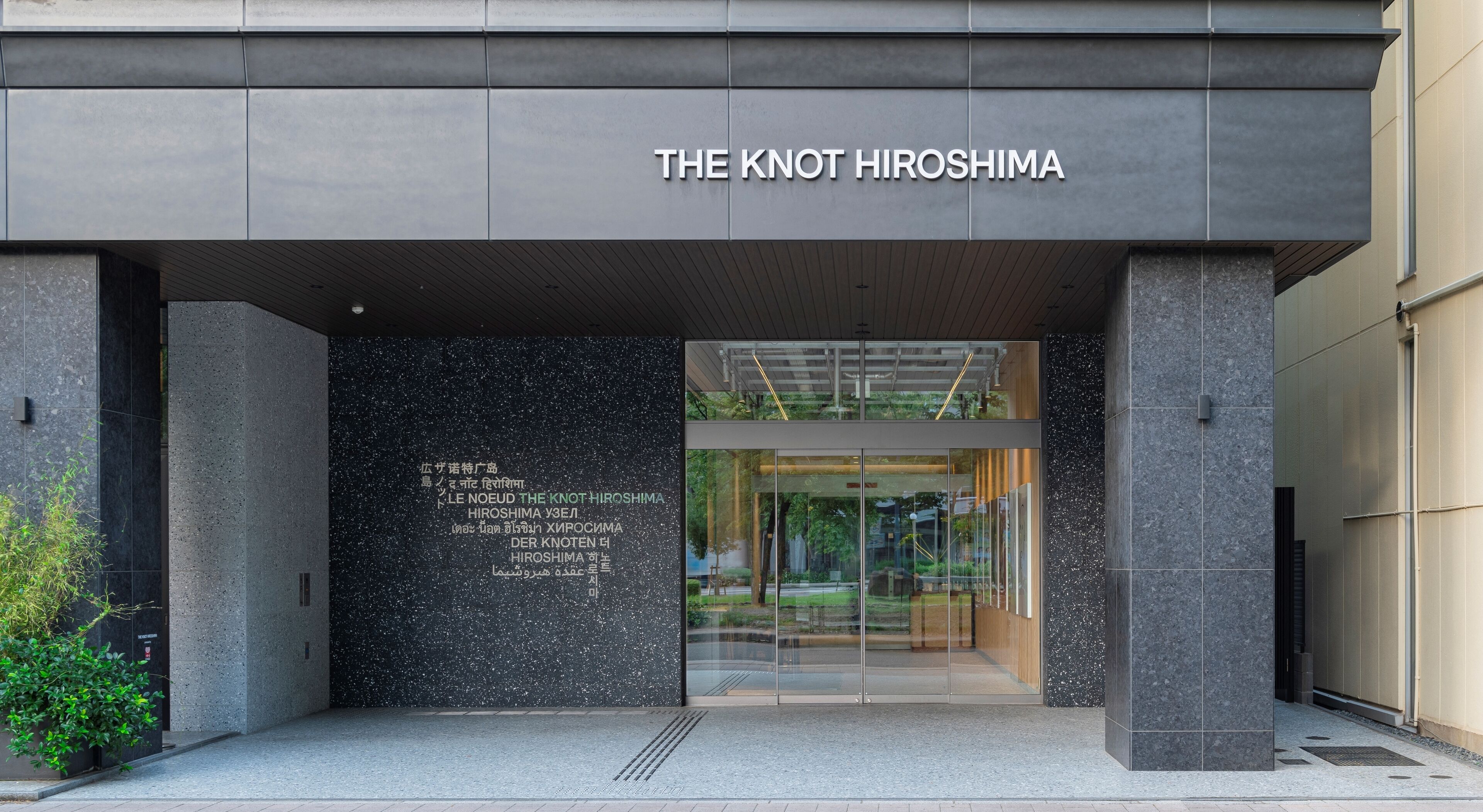 The entrance to The Knot Hiroshima with a modern design, featuring a glass door and a signboard with multiple languages.