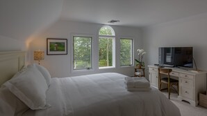 A bedroom with a bed, a desk, and a TV. There are three windows providing natural light.