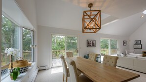 A spacious room with a large wooden dining table, comfortable seating, and large windows offering a view of the outdoors.