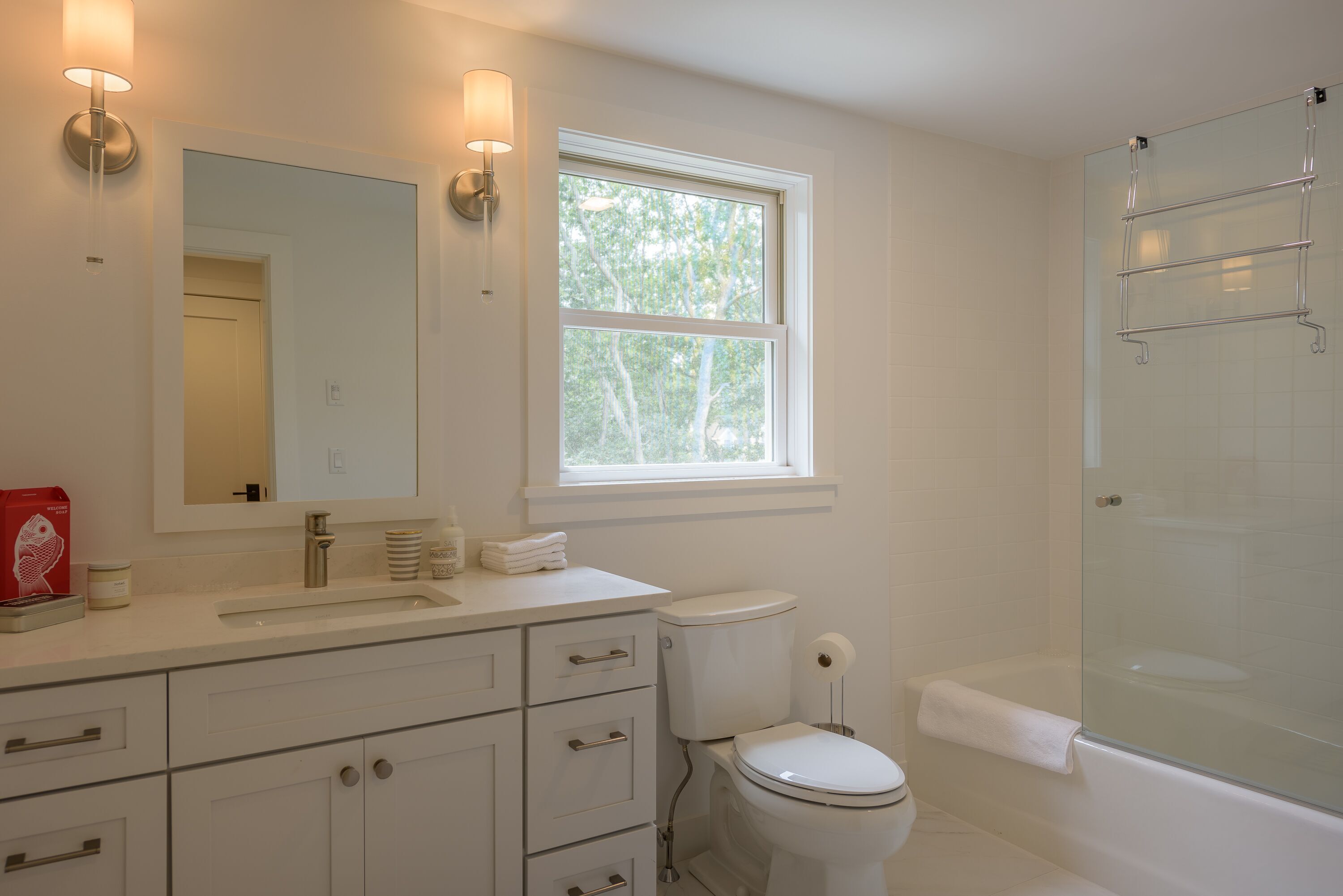 Une salle de bain moderne avec un lavabo, un miroir et des toilettes blancs, une douche en verre et une fenêtre qui laisse entrer la lumière naturelle.