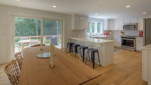 Une cuisine moderne avec des meubles blancs, un grand îlot central et un coin repas comprenant une table en bois et des chaises en osier.