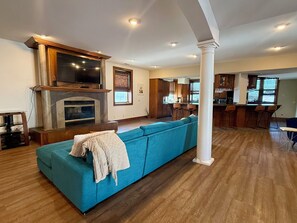 A living room with a fireplace, a large blue sofa, wooden flooring, and a kitchen area with bar stools.