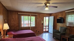 A room with two beds, a ceiling fan, a TV, and a window with a view of greenery. - The Gold Eagle Cottage 2 (Lake George)