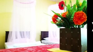Habitación de hotel con cama, mesita de noche con un jarrón de flores naranjas y ventana con cortina translúcida.