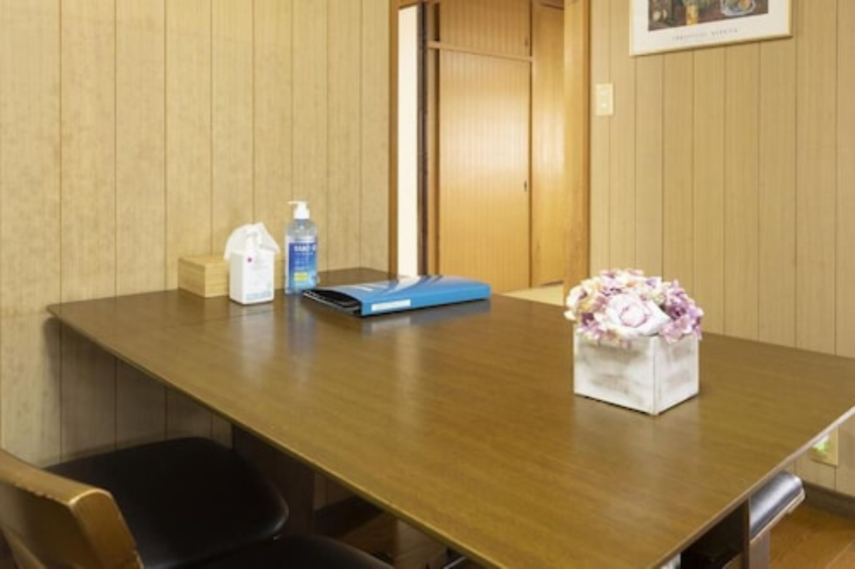 Una habitación con paneles de madera, una mesa larga, una silla, un dispensador de gel hidroalcohólico, una carpeta azul y un florero con flores.