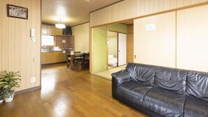 Una habitación con sofá de cuero negro, suelo de madera, una zona de cocina y una puerta corredera que da a otra habitación.