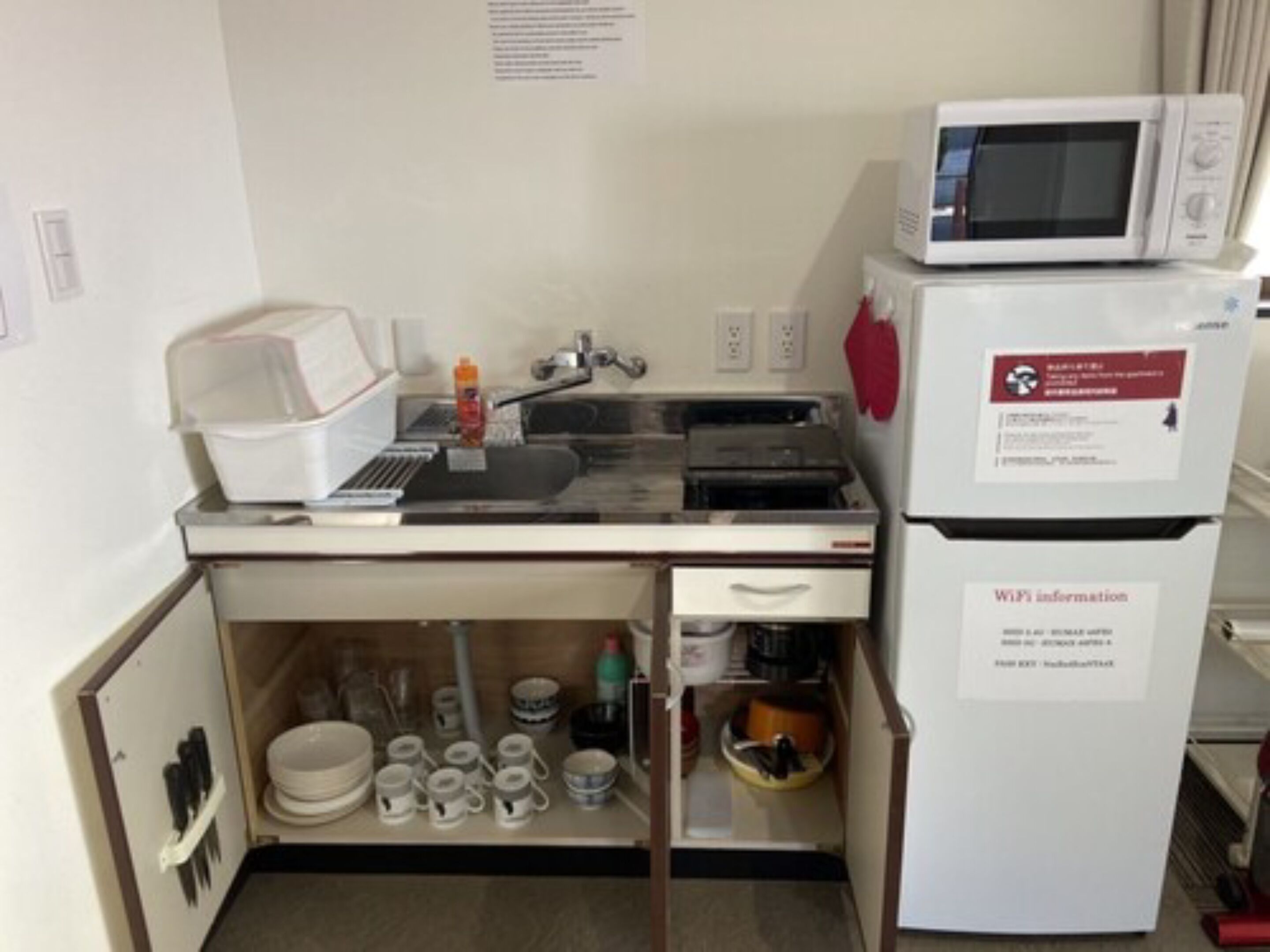 A compact kitchenette with a microwave, a small sink, and a refrigerator. There are utensils and dishes stored under the counter.