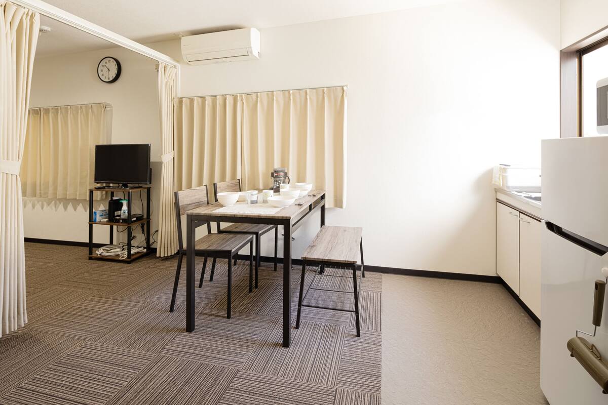 Una habitación de hotel compacta con kitchenette, un comedor con mesa y sillas, televisión y un reloj en la pared.