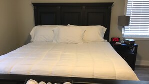 Una cama bien hecha con sábanas blancas, un cabecero oscuro y una mesita de noche con una lámpara y un reloj digital que marca las 12:06.