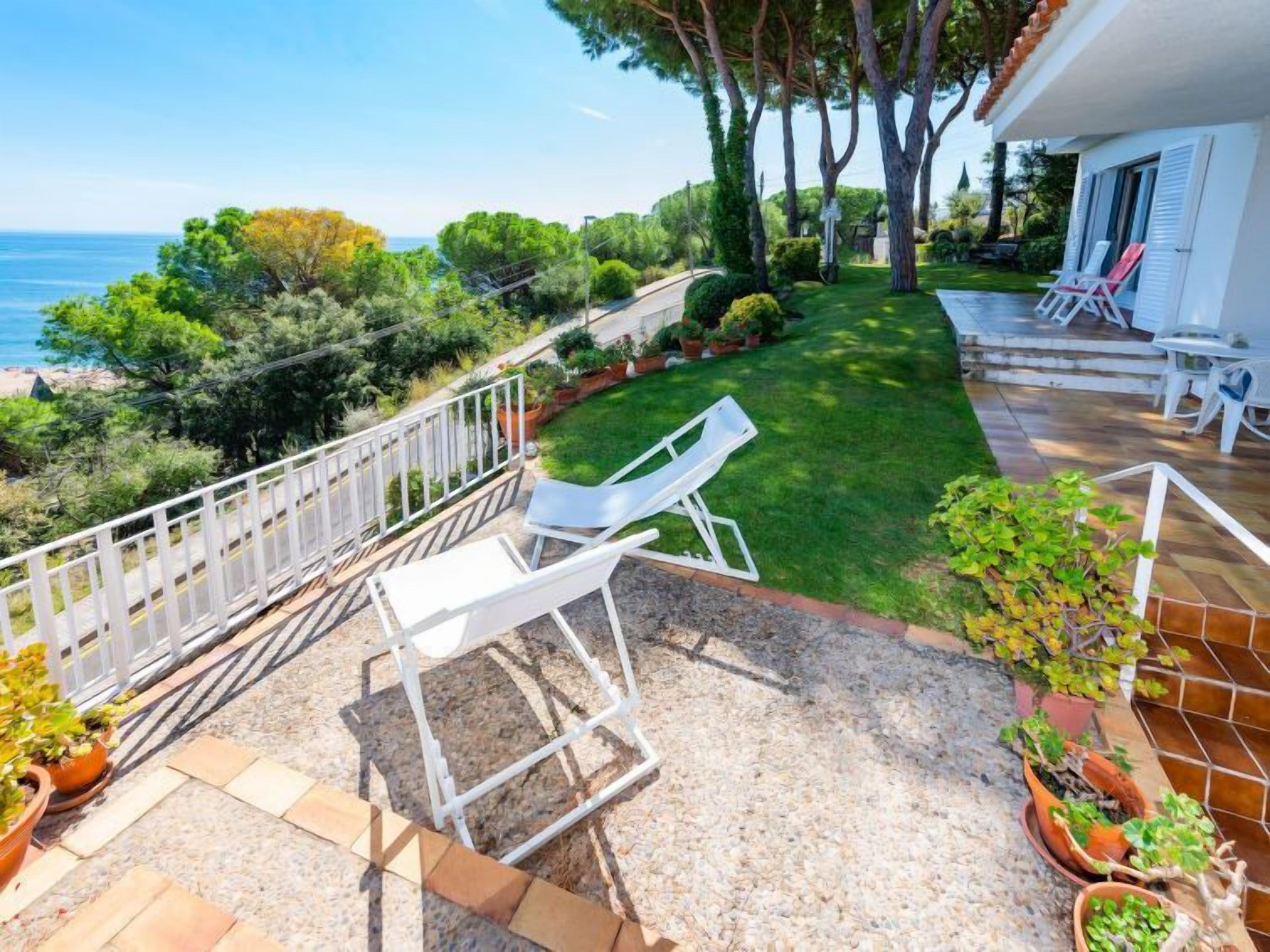 Un patio con una silla y una mesa blancas, un pequeño jardín con plantas en macetas y vistas al mar.