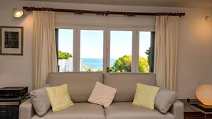 Un sofá color crema con dos almohadones, un cuadro enmarcado en la pared y vista al mar a través de amplios ventanales.