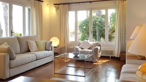 Una sala de estar con sofá, sillón, mesa de centro y ventanas grandes con cortinas.