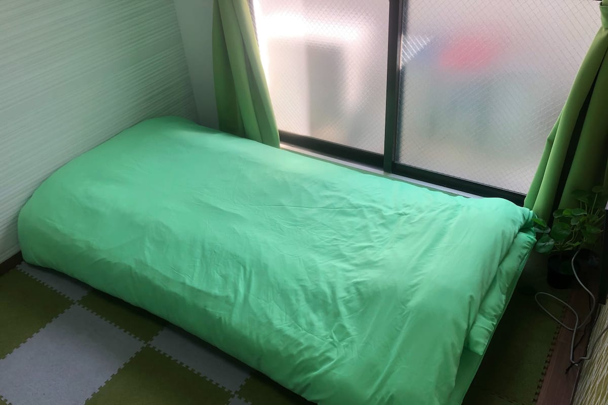 A small, single-bed room with a green bedspread, a window with green curtains, and a potted plant.