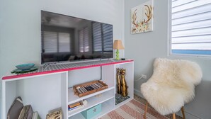 A living room with a flat-screen TV on a white entertainment unit, a fluffy chair, and a window with blinds. - The Babanees - Bummer Hill Boho Chic Surf Shack  (Rincon)