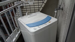 Eine kompakte, weiße Waschmaschine mit blauem Bedienfeld, in einem schmalen Raum mit Metallgeländer und gefliesten Wänden.