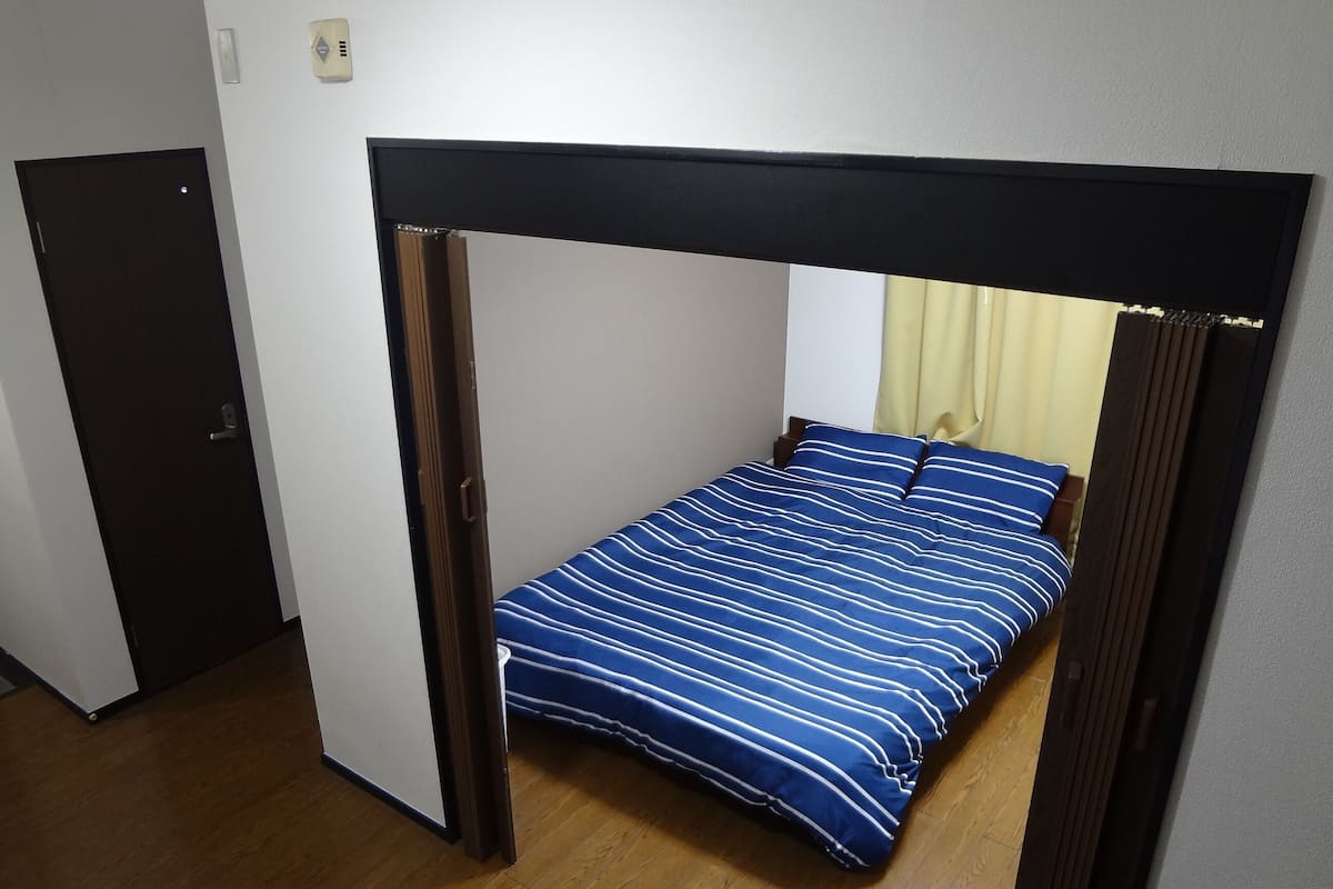 Una cama individual con cabecera oscura y colcha a rayas azules en una habitación con suelo de madera y una puerta cerrada.