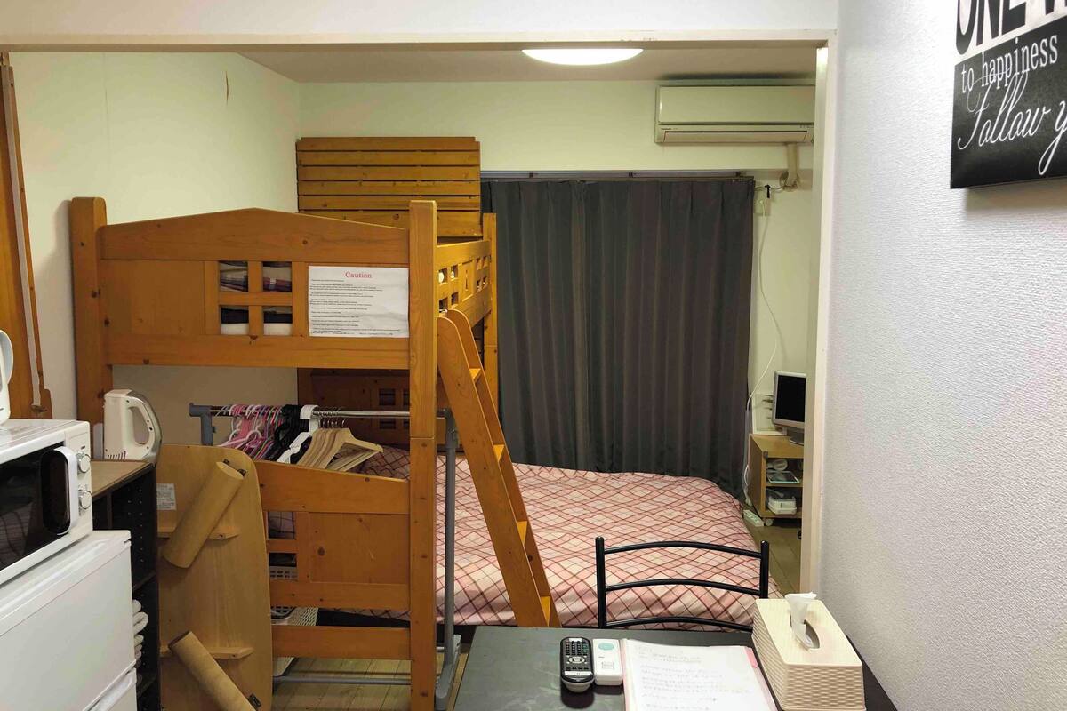 Una habitación pequeña y funcional con una cama litera, un escritorio con computadora, una silla, una mesita y un televisor instalado en la pared.