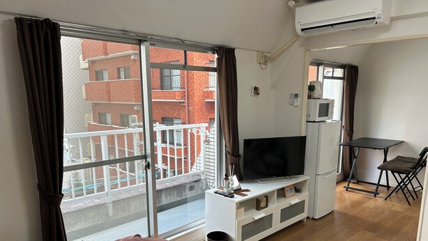 A modern hotel room with a large sliding glass door to a balcony, a flat-screen TV on a white entertainment unit, a small table with a chair, and a refrigerator. - Guest House Zen Hakata Ohori Park 2 (Fukuoka)