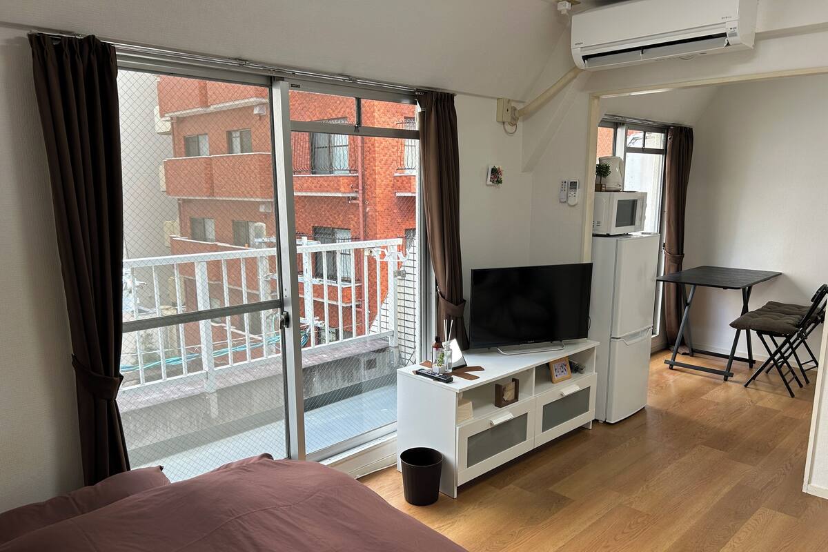 Une chambre d’hôtel moderne dotée d’une grande porte-fenêtre coulissante donnant sur un balcon, d’un téléviseur à écran plat installé sur un meuble blanc, d’une petite table avec une chaise et d’un réfrigérateur.