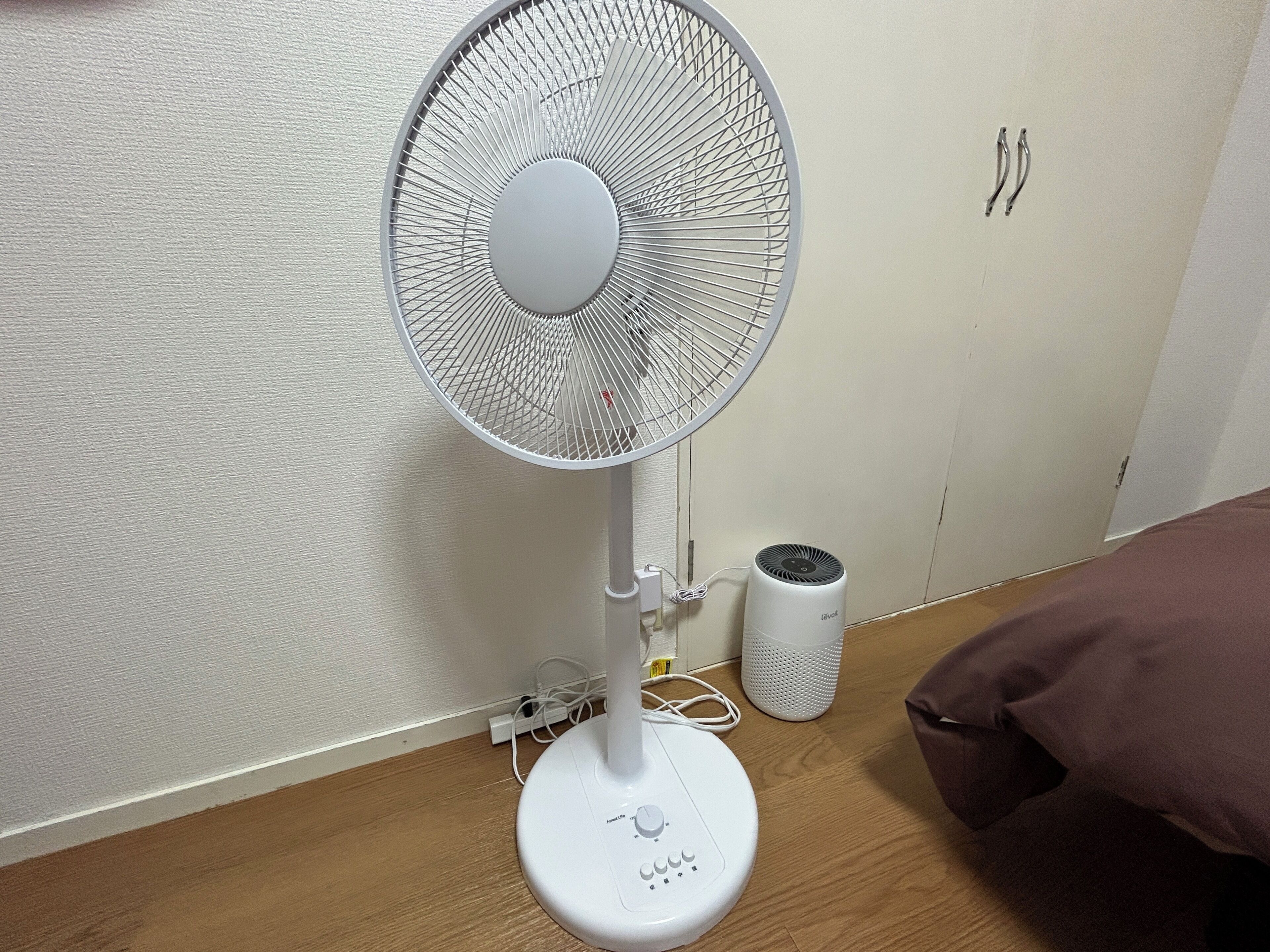 A white standing fan with a control panel, a white cylindrical device, and a brown pillow on a wooden floor.