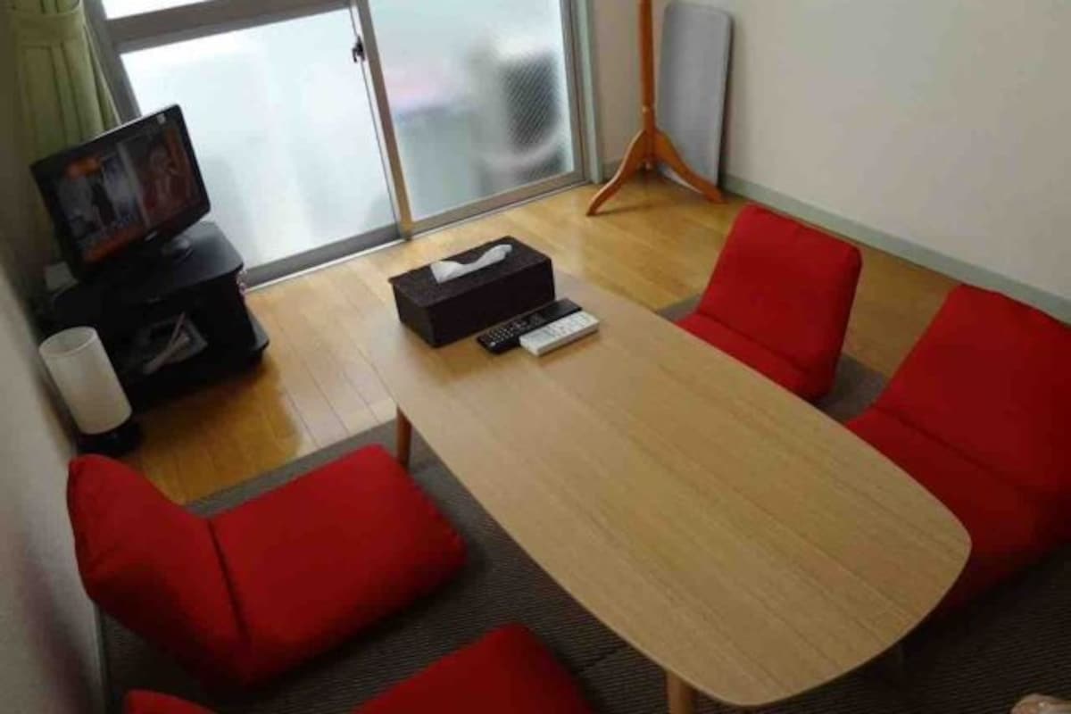 Een vergaderruimte met een lange tafel en vier rode stoelen, een televisie op een standaard en een doos met een afstandsbediening.