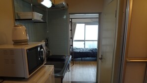 Een kleine keuken met een magnetron, waterkoker en wastafel. Een bed is zichtbaar door een open deur, die leidt naar een kamer met een raam.