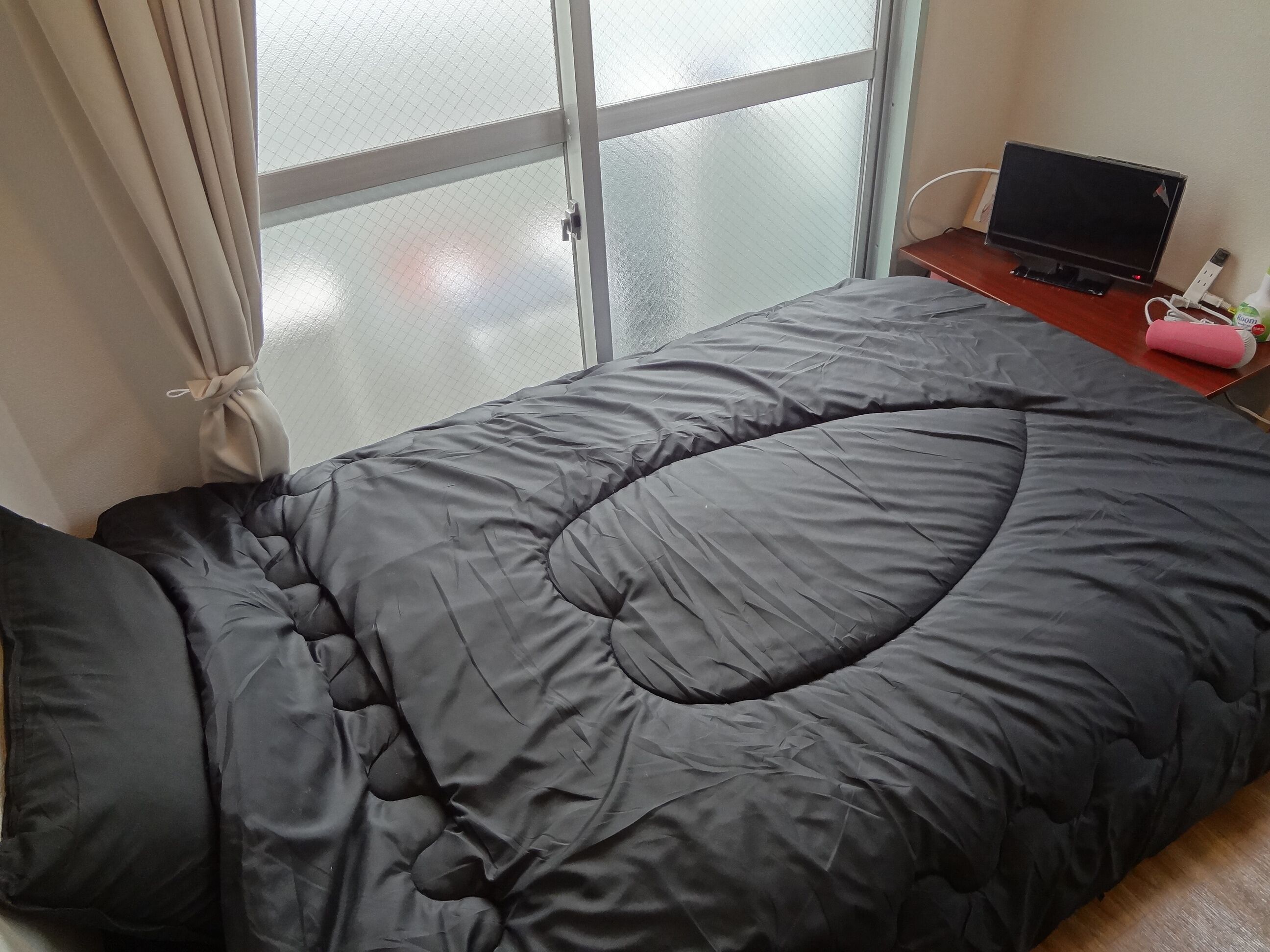 Habitació amb un llit cobert per un edredó gris, una televisió sobre una taula de fusta i una finestra amb cortines.