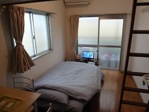 A bedroom with a bed, a desk, a chair, a TV on a stand, and a window with curtains.