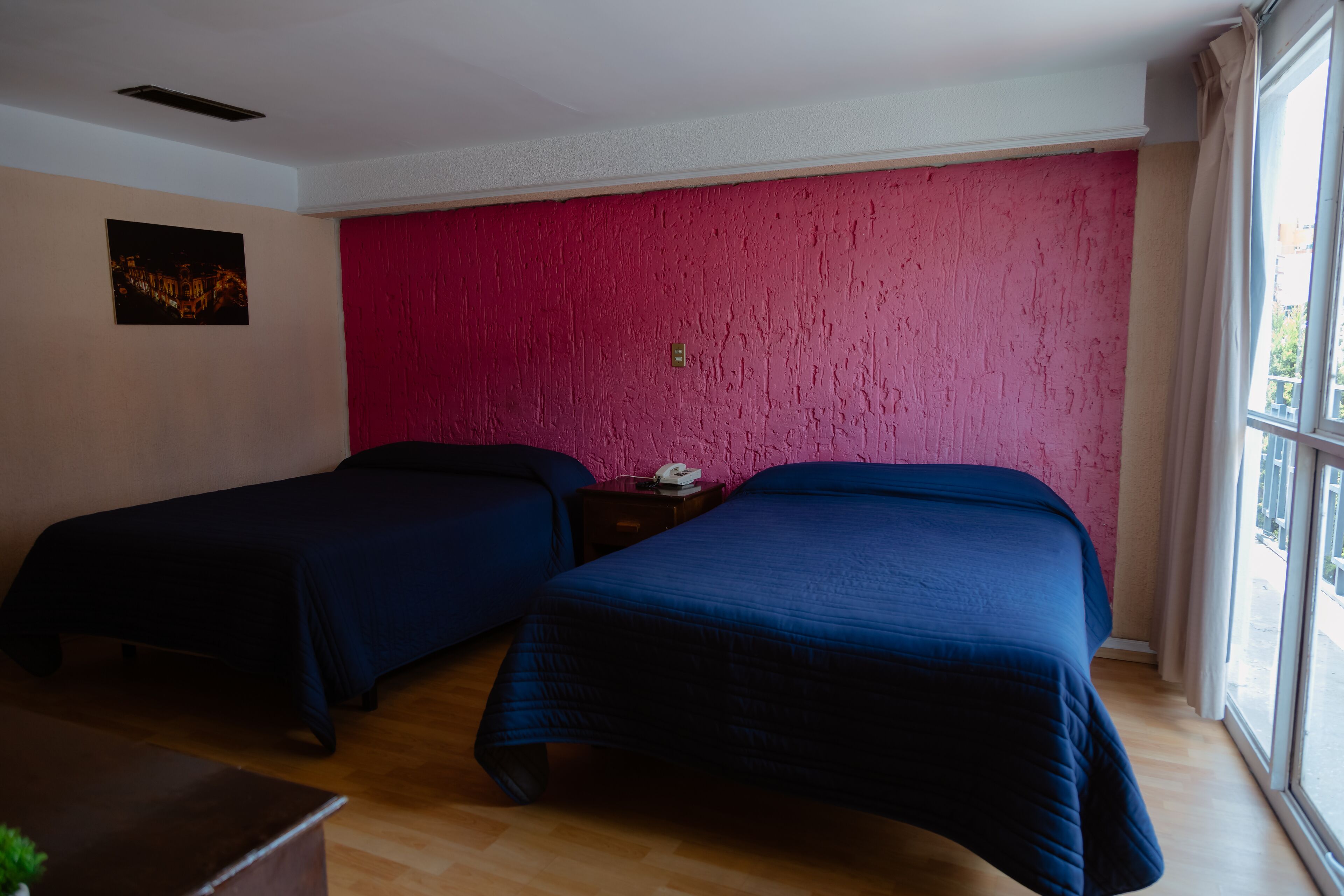 Una habitación con dos camas, suelo de madera, pared rosa con textura y una ventana con vistas.