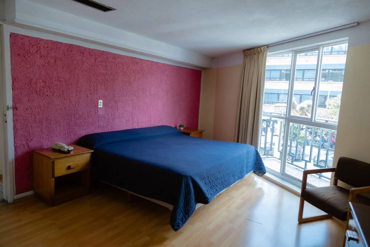 Ein Zimmer mit einem Bett, Nachttischen aus Holz, einem Sessel, einem Fenster mit Blick nach draußen und einer Wand mit rosa Struktur.