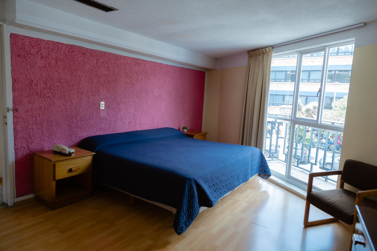 Ein Schlafzimmer mit einem großen Bett, Nachttischen aus Holz, einem Sessel, einem Fenster mit Blick ins Freie und einer Wand in Rosé mit Struktur.