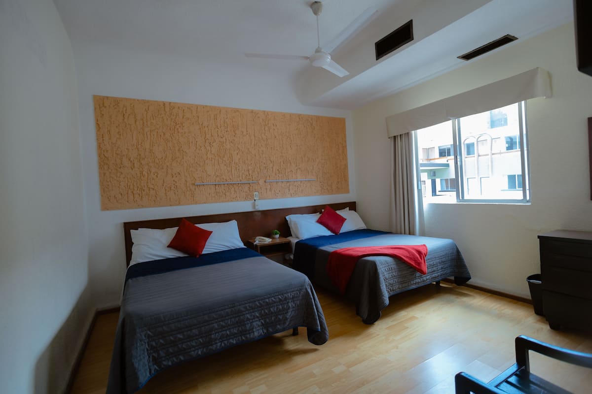 Ein Hotelzimmer mit zwei Betten, einem Deckenventilator, einem Fenster mit Blick auf Gebäude und einem Holzboden.