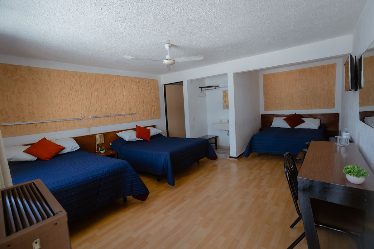 Ein Hotelzimmer mit drei Betten, Holzboden, Deckenventilator, Schreibtisch mit Stuhl und einer kleinen Pflanze auf dem Schreibtisch.