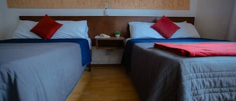 A hotel room with two beds, each with a blue bedspread and red pillows. There is a small bedside table with a plant and a phone.
