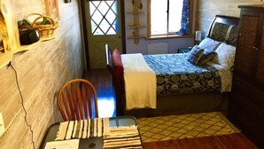 A small, cozy bedroom with a single bed, a chair, a desk with a computer, a window with curtains, and a door with a cross-shaped window. - Small studio in the quite neighbourhood. (Flagstaff)
