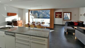 Une cuisine moderne avec un coin repas, de grandes fenêtres offrant une vue sur les arbres et les montagnes, et un salon avec un canapé et un tableau.