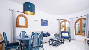 Una sala de estar moderna con una mesa redonda de comedor, sillas azules, un sofá y una mesa de centro. Hay dos ventanas grandes con marcos de madera y cortinas blancas.