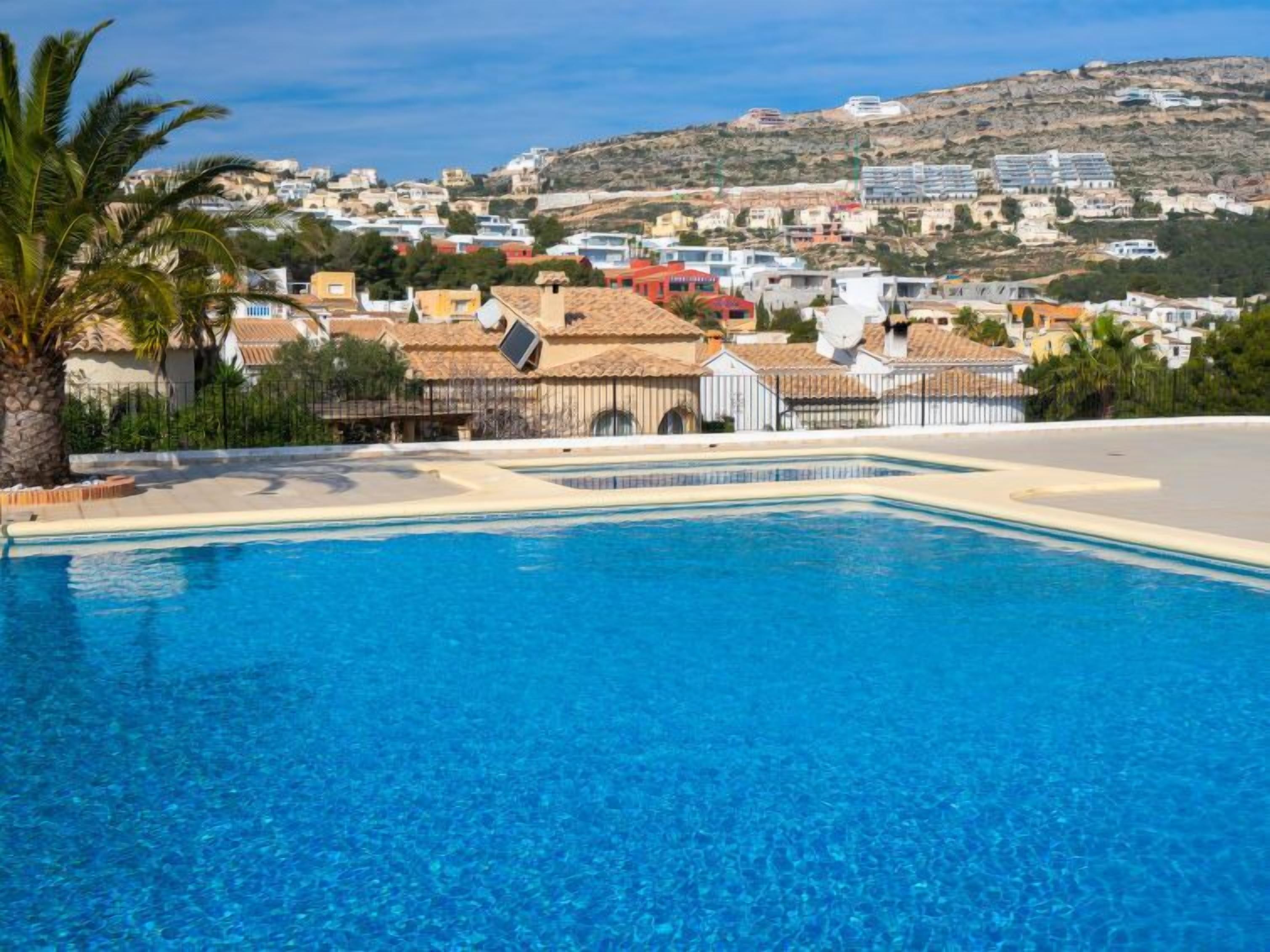 En swimmingpool med klart blåt vand, omgivet af bygninger i middelhavsstil, med terracotta-tage og hvide vægge.