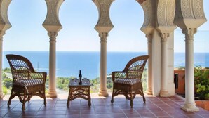 A covered outdoor seating area with three wicker chairs and a small table, overlooking a scenic view of the sea.