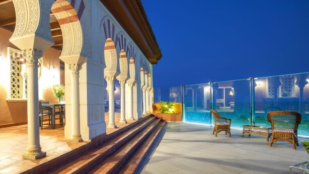 A rooftop pool area with steps leading down to a bar area, surrounded by ornate arches and columns.