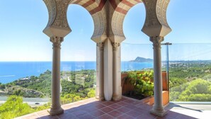 A covered outdoor area with arches and columns, overlooking a scenic landscape with greenery and a view of the sea.