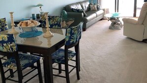 A dining area with a table set for four, surrounded by chairs with floral upholstery. A sofa and a chair are positioned against the wall, with a lamp on a side table. - Condo with 2 bedrooms (Jensen Beach)
