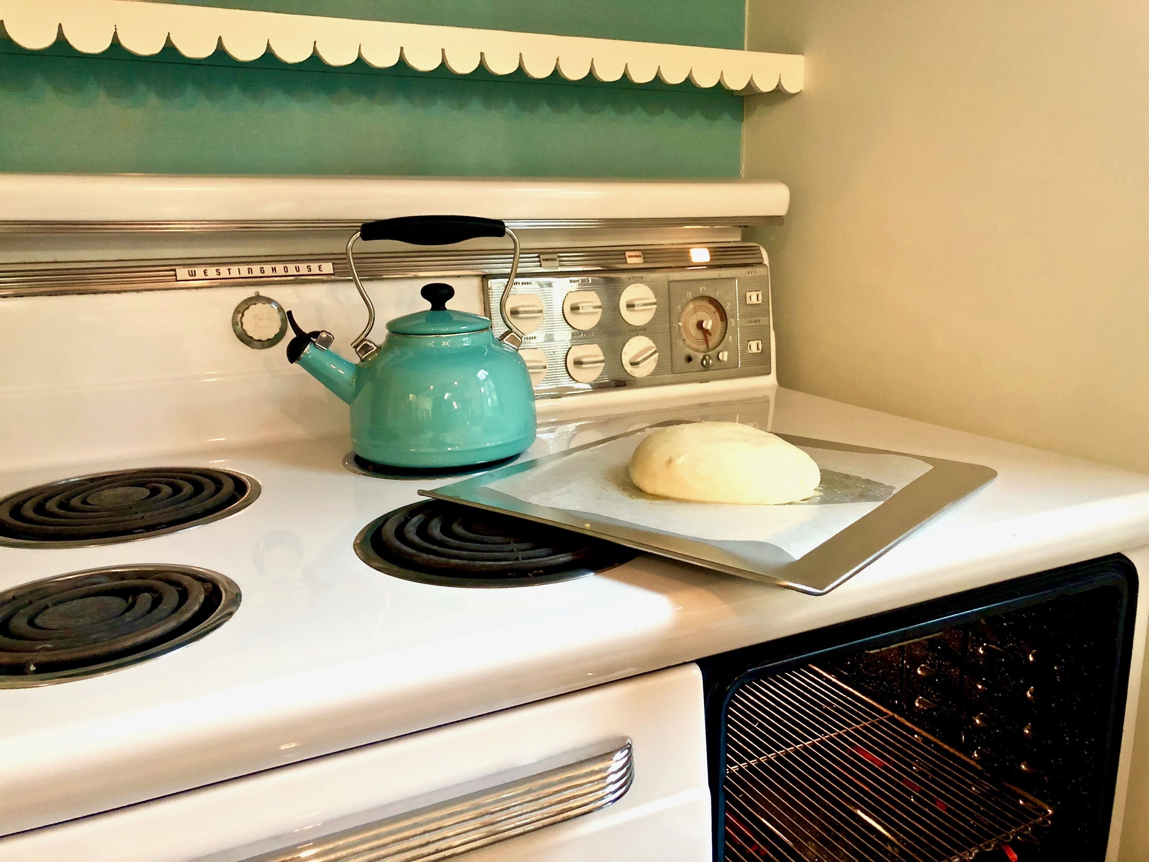 A kitchen with a white stove, a teal kettle, and a loaf of bread on a baking tray.